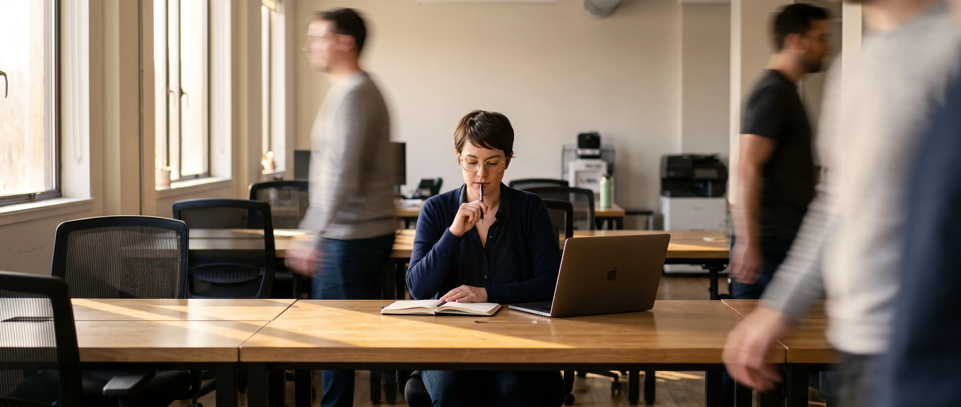 An engineer thinks over her notebook at a sunlit shared desk while colleagues walk past
